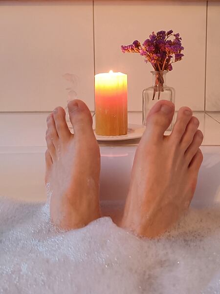 Relaxace ve vaně, pohled je na chodidla, která vykukují z pěny. Mezi chodidly je na vaně postavena svíčka, která umocňuje relaxaci.