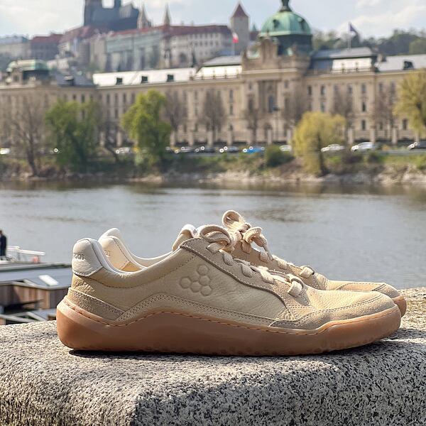 Pohled na Úřad vlády ČR a Pražský hrad, které jsou vidět v dálce. Detail je na boty Vivobarefoot. Jedná se o městské tenisky v béžové barvě s bílým detailem na patě.