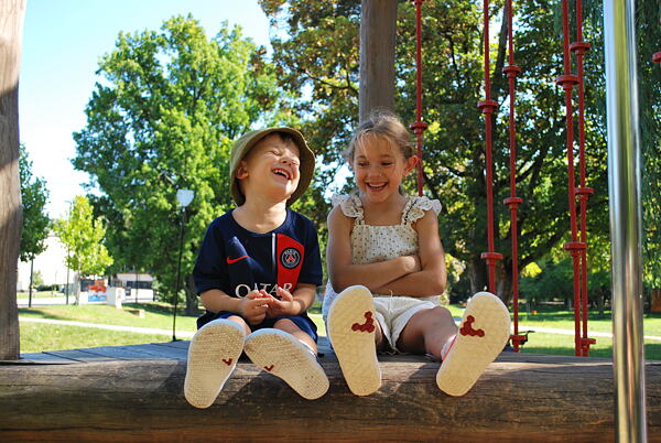 Smějící se dvě děti - chlapec a dívka - které sedí na dětském hřišti. Na nohou mají boty VIVOBAREFOOT. 