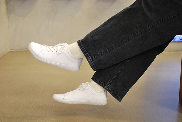 Na fotce je pohled na nohy člověka, který má černé džíny, světlé ponožky a k tomu bílé boty VIVOBAREFOOT Geo Court.